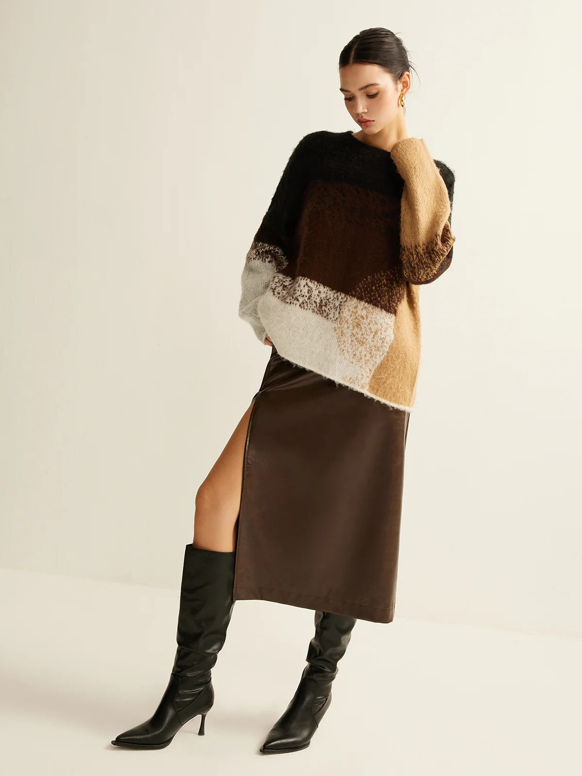 Woman wearing a brown and beige sweater with a slit skirt and black knee-high boots on a plain background