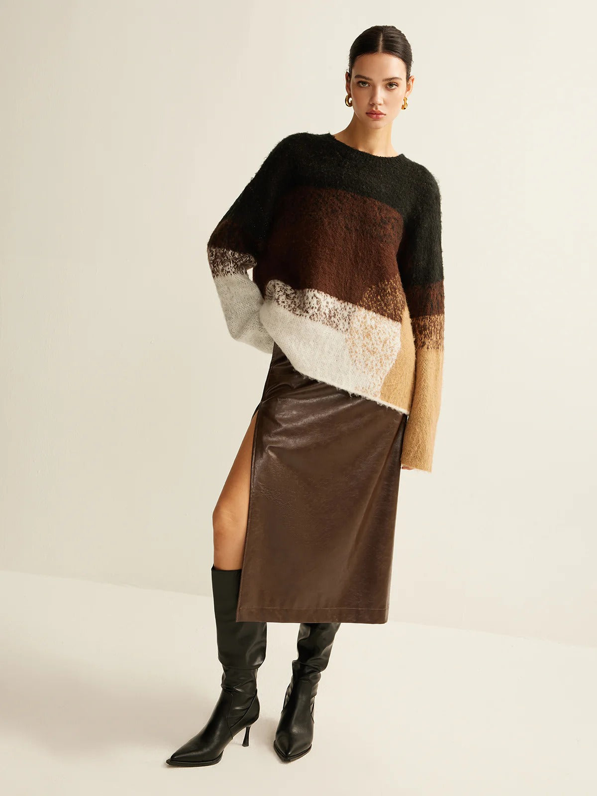 Woman wearing a brown and black sweater with a white pattern, brown leather skirt, and black knee-high boots on a light background.