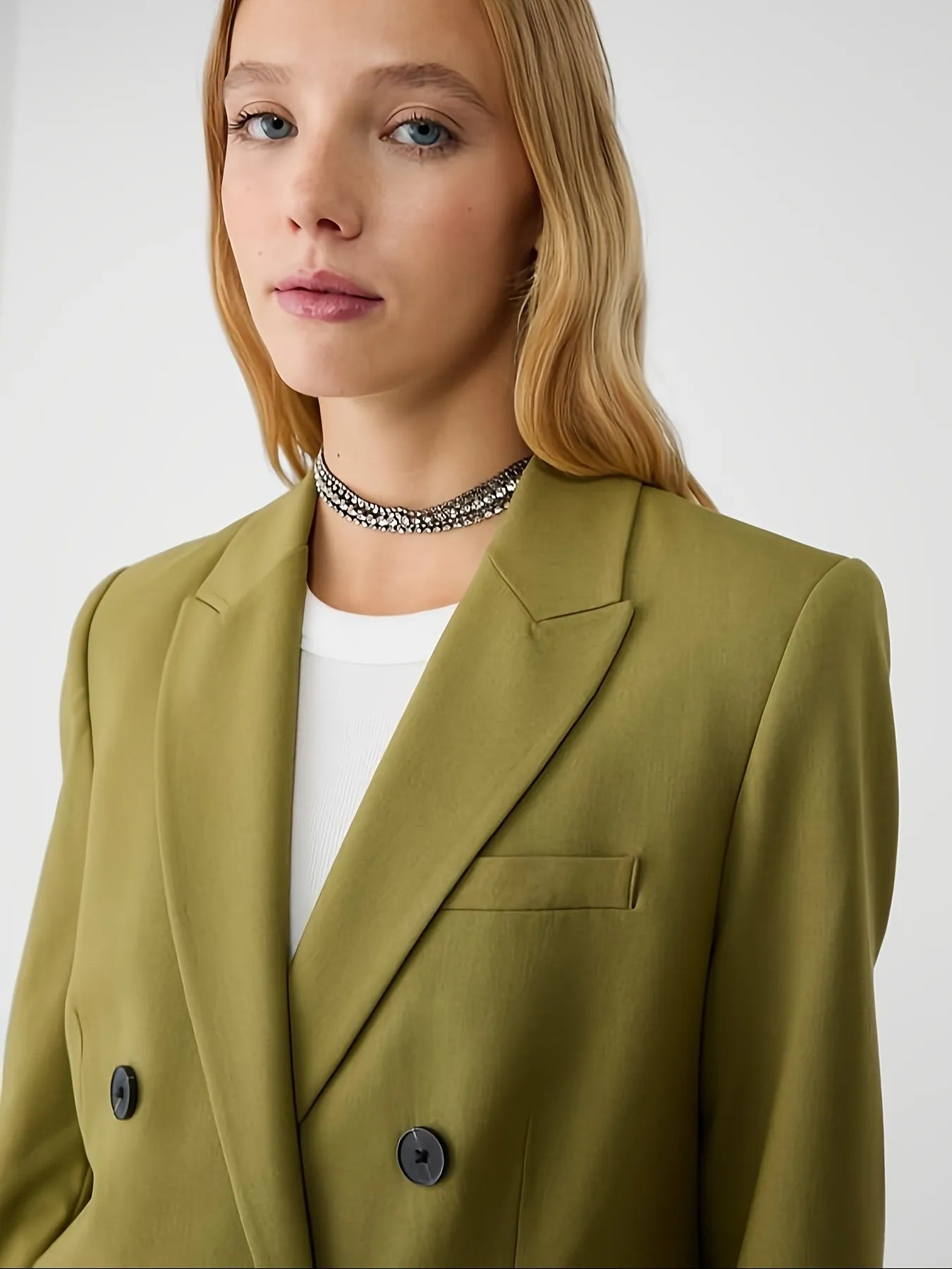 Woman wearing a green jacket with a tailored fit, layered over a cream long-sleeve top with black contrast wavy stitching, paired with high-waisted blue jeans. The look is completed with hoop earrings and a sleek bun hairstyle for a modern minimalist aesthetic.
