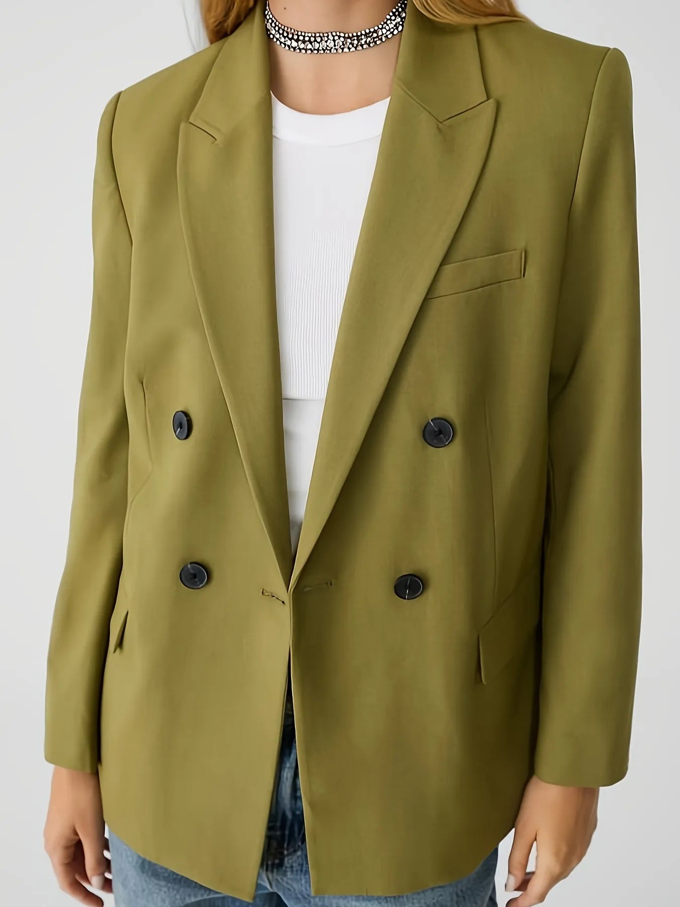 Woman wearing a green jacket with a tailored fit, layered over a cream long-sleeve top with black contrast wavy stitching, paired with high-waisted blue jeans. The look is completed with hoop earrings and a sleek bun hairstyle for a modern minimalist aesthetic.