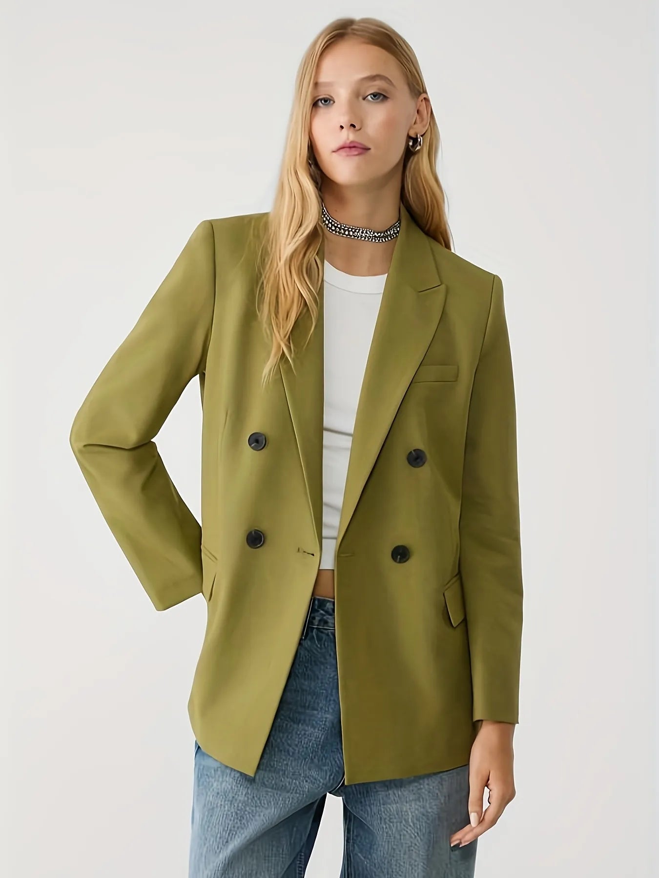 Woman wearing a green jacket with a tailored fit, layered over a cream long-sleeve top with black contrast wavy stitching, paired with high-waisted blue jeans. The look is completed with hoop earrings and a sleek bun hairstyle for a modern minimalist aesthetic.