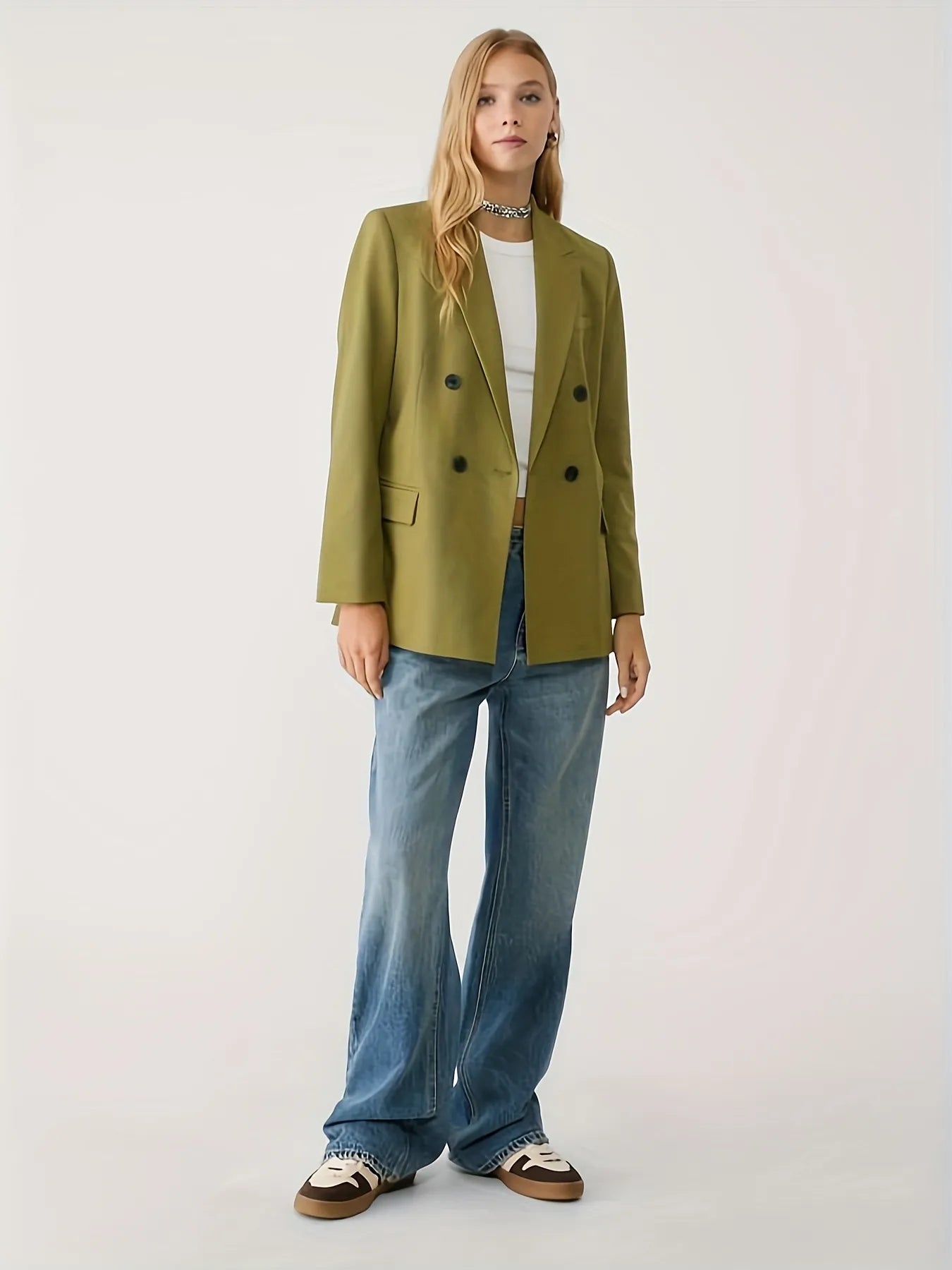 Woman wearing a green jacket with a tailored fit, layered over a cream long-sleeve top with black contrast wavy stitching, paired with high-waisted blue jeans. The look is completed with hoop earrings and a sleek bun hairstyle for a modern minimalist aesthetic.