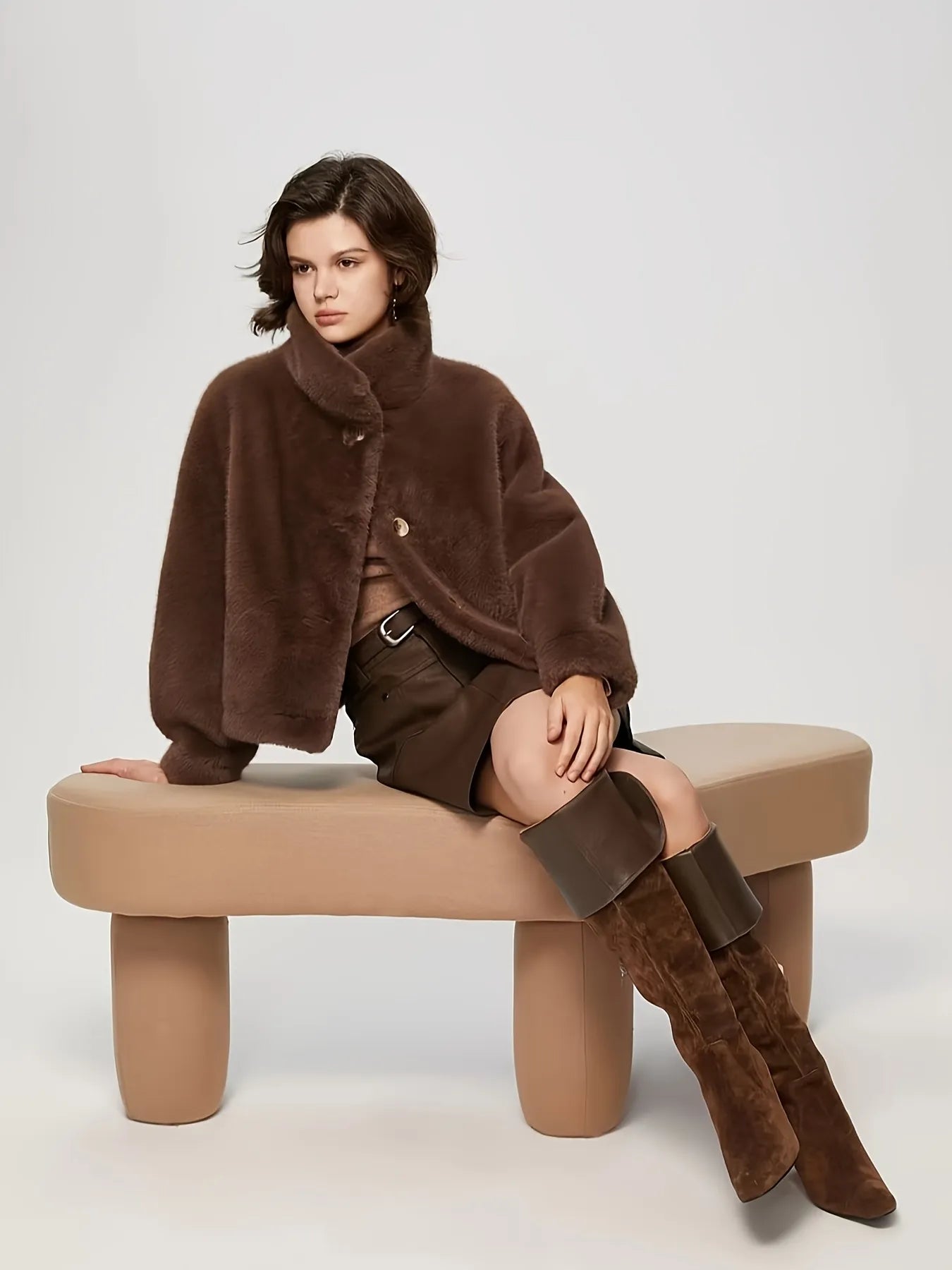Woman wearing a brown fur coat and boots sitting on a beige bench against a white background