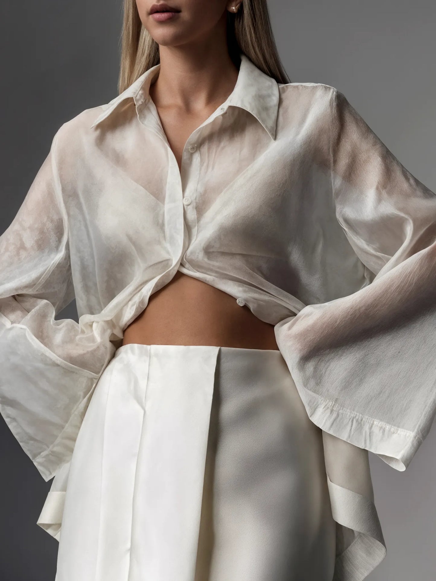 Person wearing a light-colored sheer shirt over a white outfit against a gray background