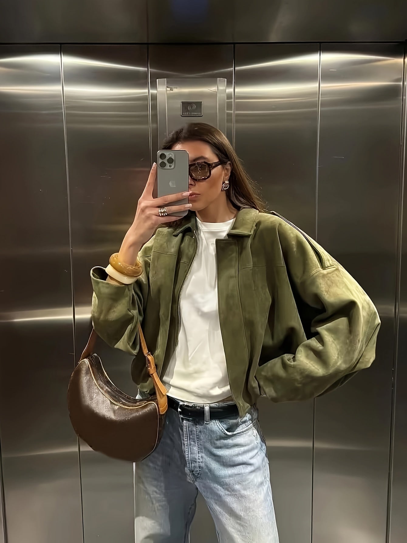 Person taking a mirror selfie wearing a green jacket, white shirt, blue jeans, and brown bag in an elevator.