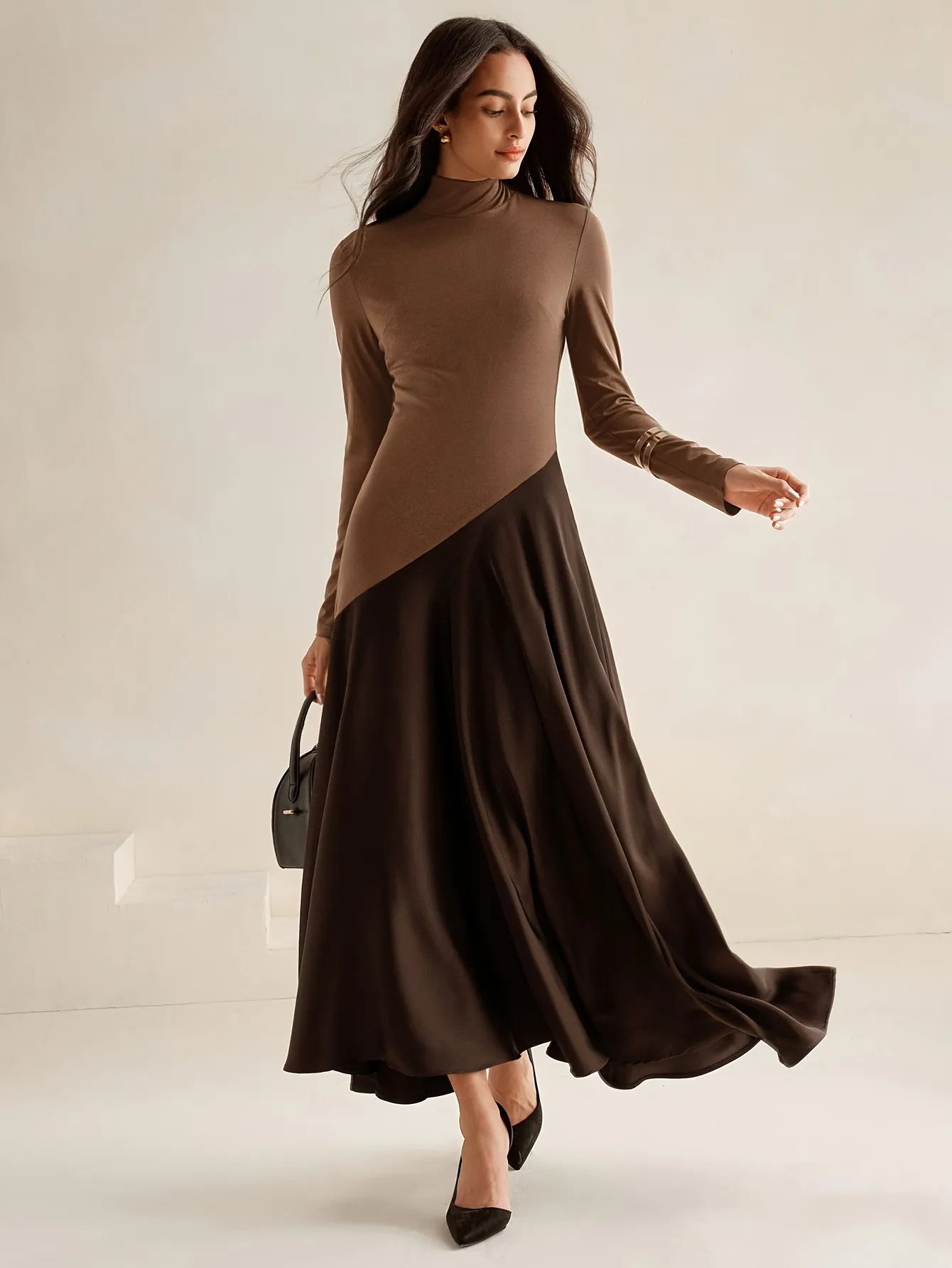 Model wearing a long-sleeve brown high-neck top dress with a diagonal two-tone design and flowing dark brown maxi skirt, paired with black pointed heels, styled in an elegant minimalist look.