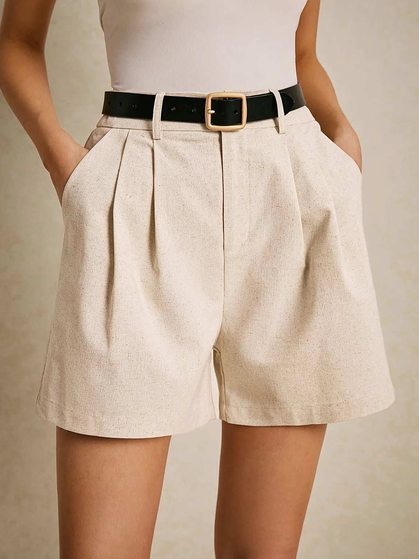 Woman wearing a white thin-strap top, high-waist beige pleated shorts with a black belt, in a smart-casual style.
