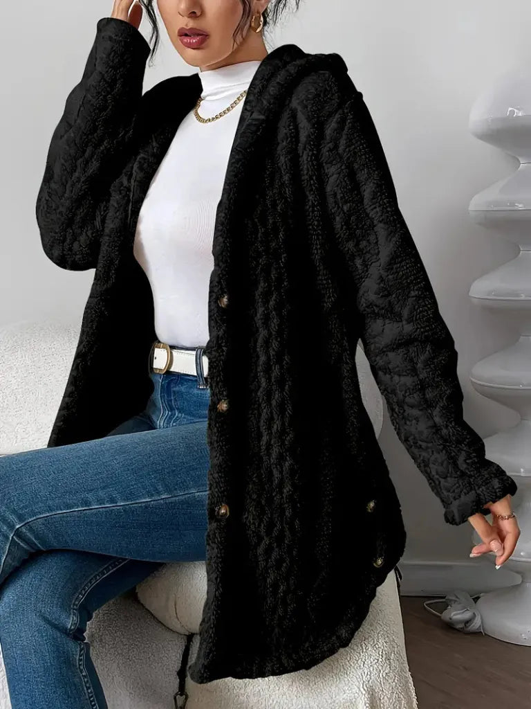 Person wearing a black textured coat over a white top and blue jeans, sitting on a white surface.