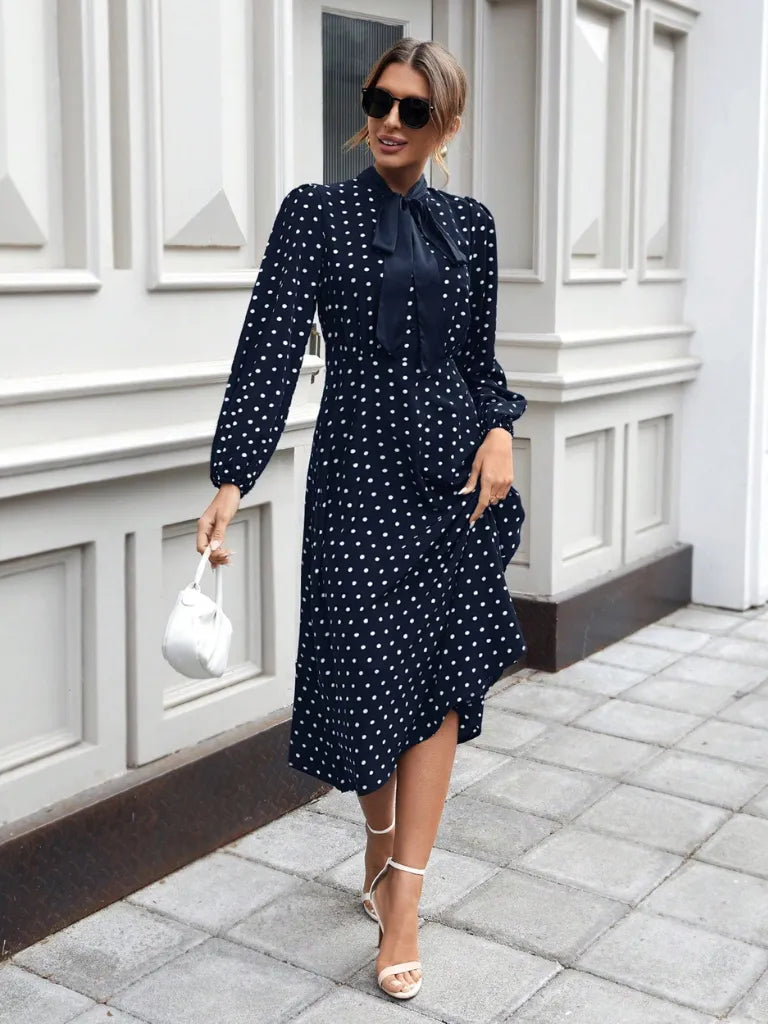 Woman wearing a navy polka dot dress standing on a sidewalk.