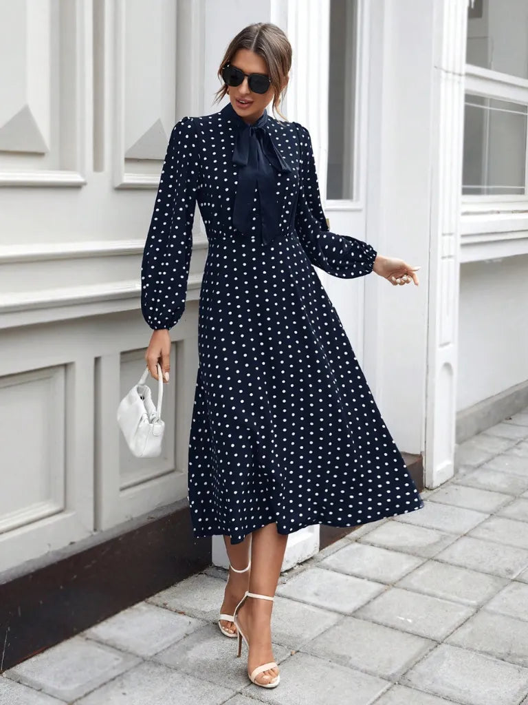 Woman wearing a navy polka dot dress with white heels and sunglasses, standing on a sidewalk.
