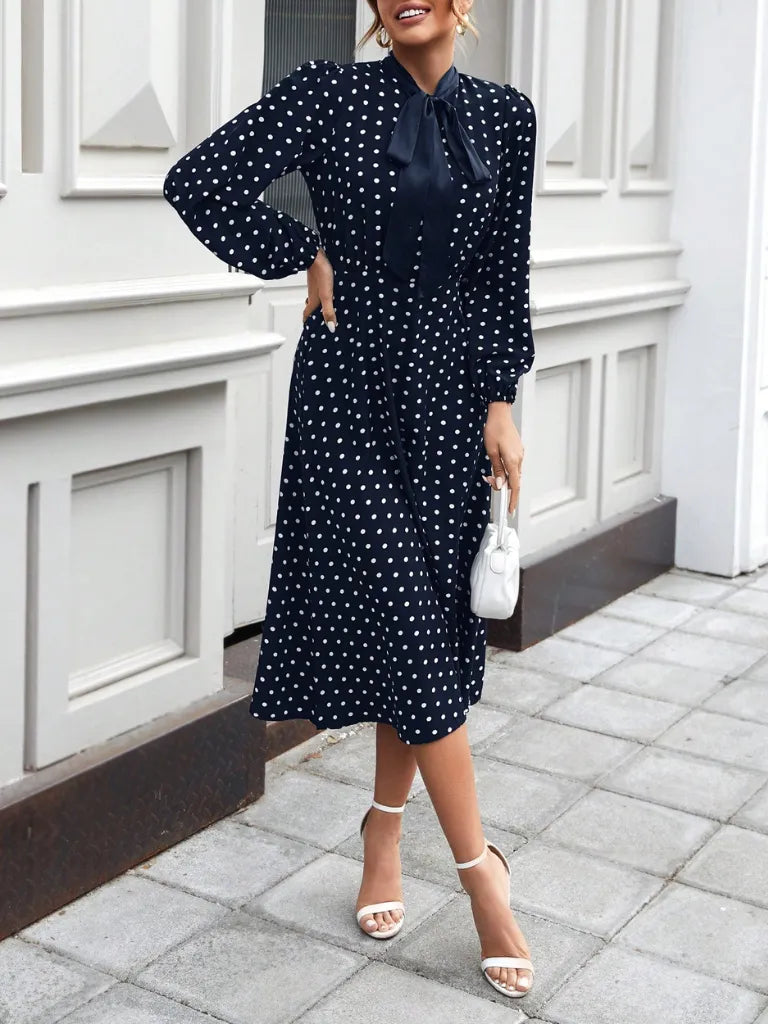 Woman wearing a navy blue dress with white polka dots, standing on a sidewalk.