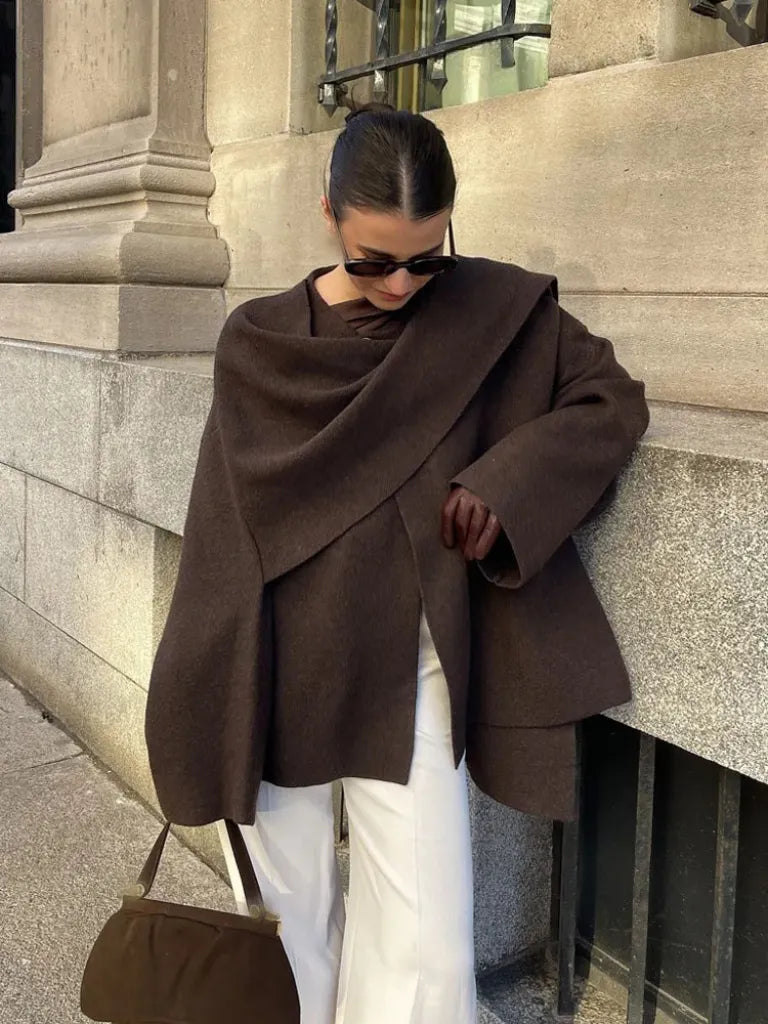 Person wearing a brown coat and sunglasses, leaning against a stone wall.