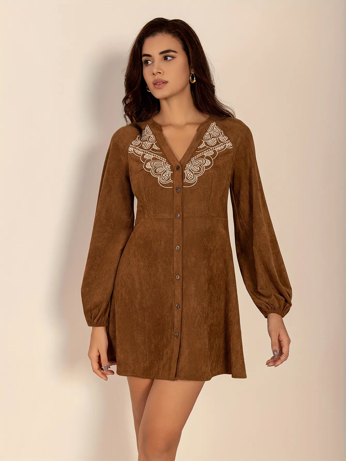 Woman wearing a short brown button-up dress with long puffed sleeves and embroidered lace detail on the chest. The dress has a relaxed fit and a soft suede-like texture, styled with simple gold hoop earrings.