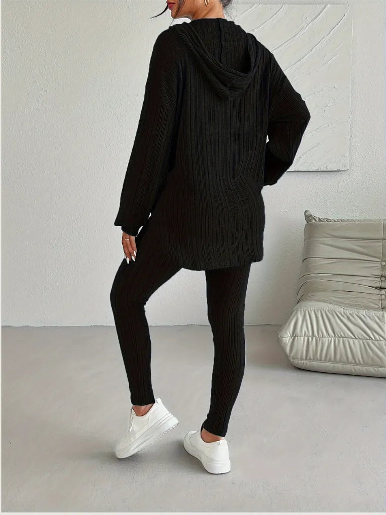 Person wearing a black knit hoodie and leggings in a minimalistic room.