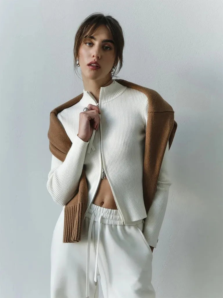 Woman wearing a white ribbed sweater with a brown leather jacket over it, on a plain background.