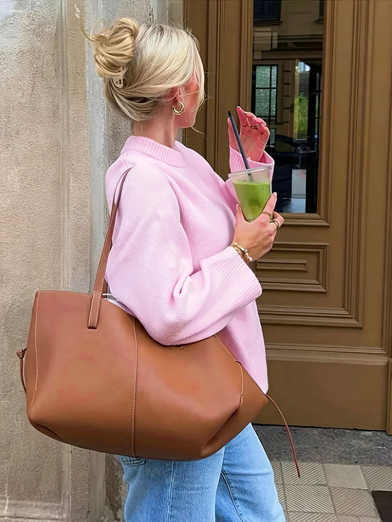 Woman in pink sweater holding a green drink and brown bag, standing in front of a door.