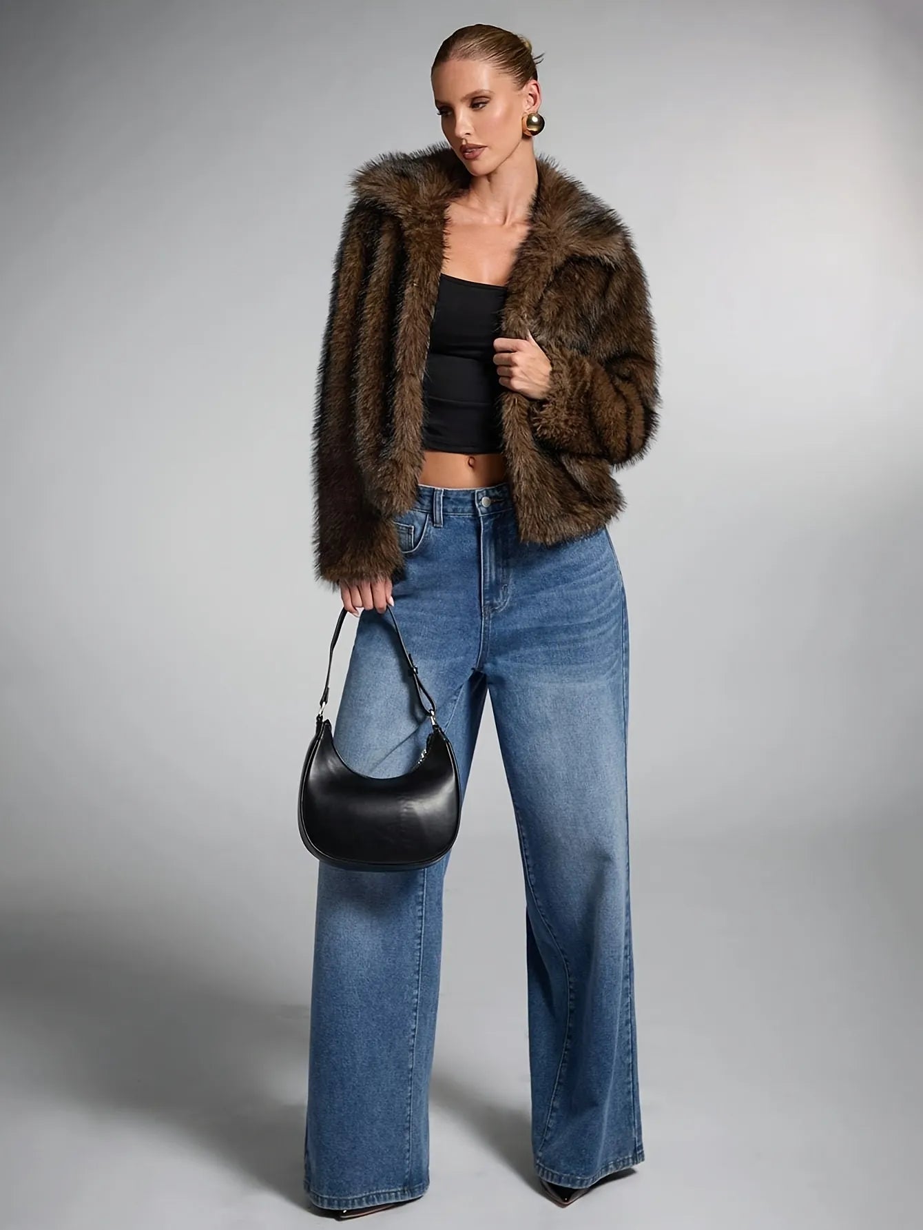 Woman wearing a fur coat, black top, and blue jeans holding a black handbag on a gray background