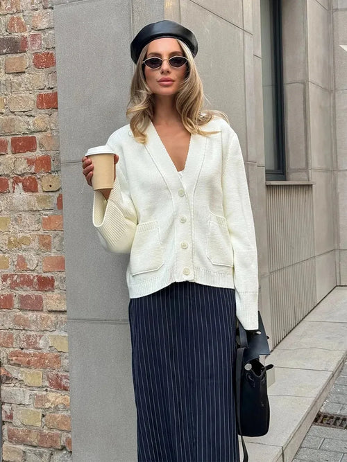 Woman in a white cardigan and navy pinstripe pants holding a coffee cup against a brick wall.