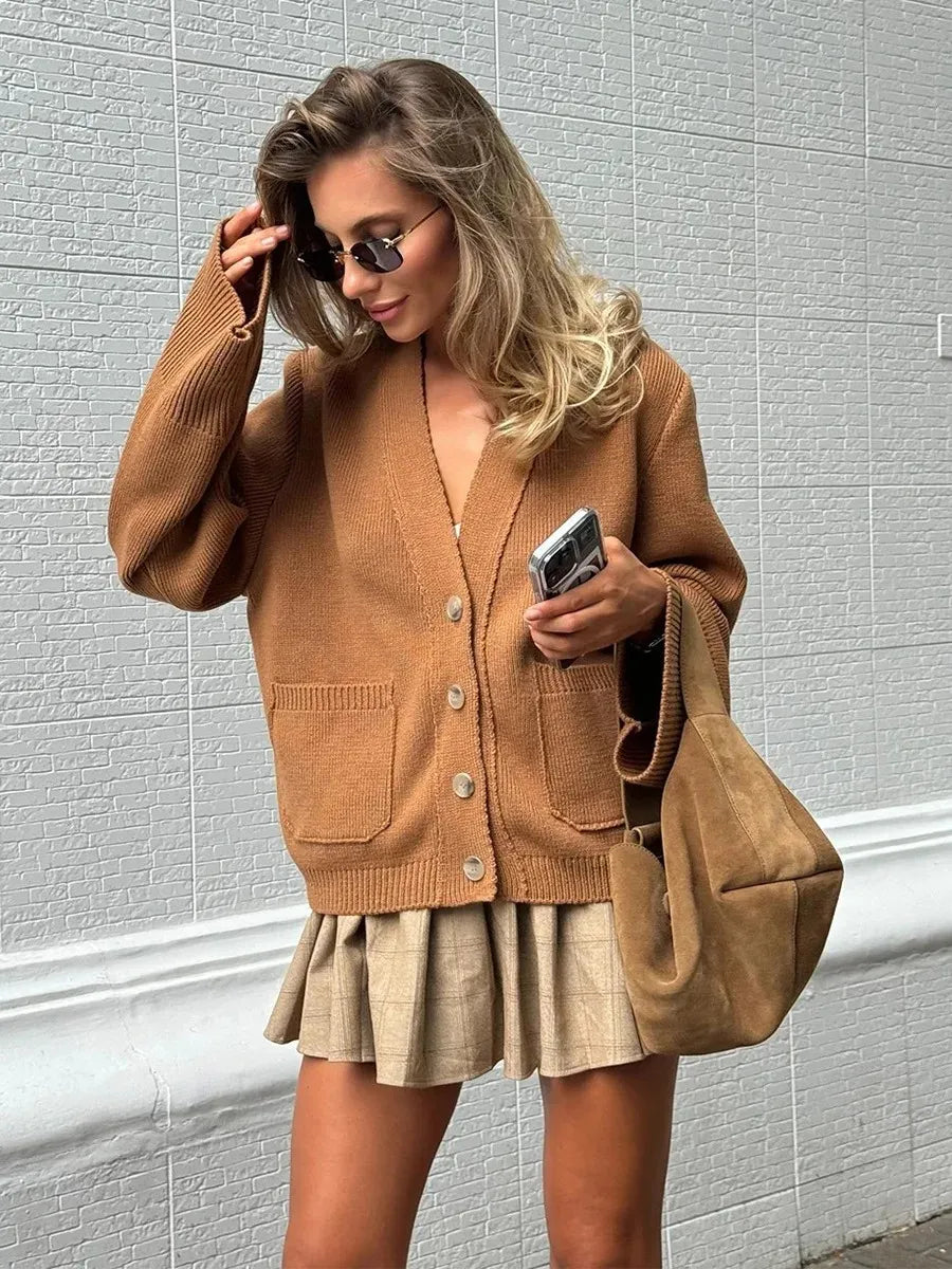 Woman wearing a brown cardigan and skirt with a beige bag against a light gray wall.