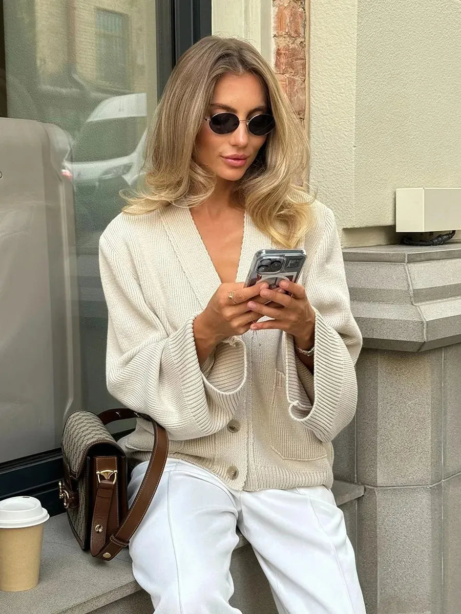 Woman in a beige cardigan and white pants sitting on a step, looking at her phone.