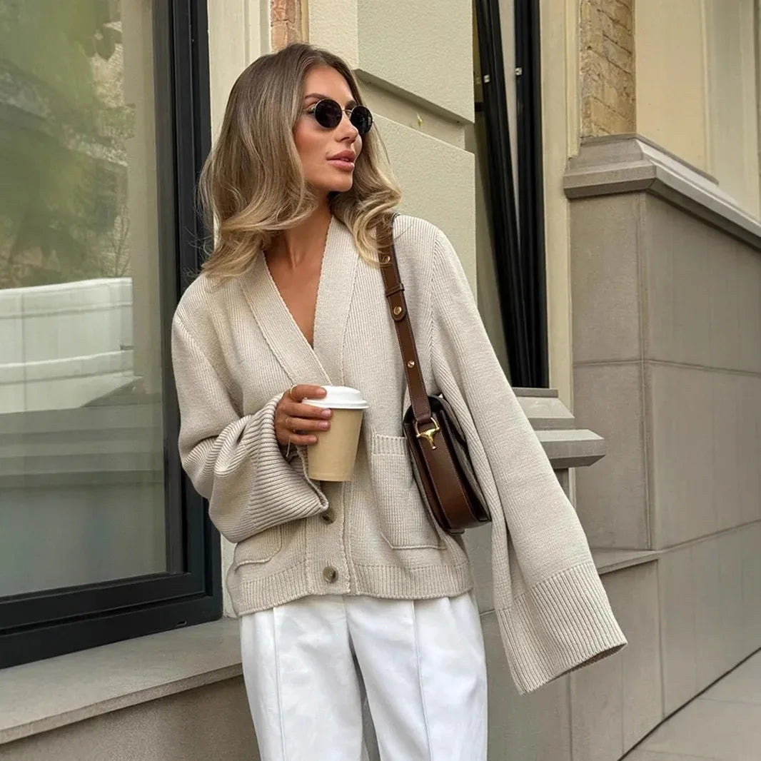 Woman in beige cardigan and white pants holding a coffee cup, standing outside a building.
