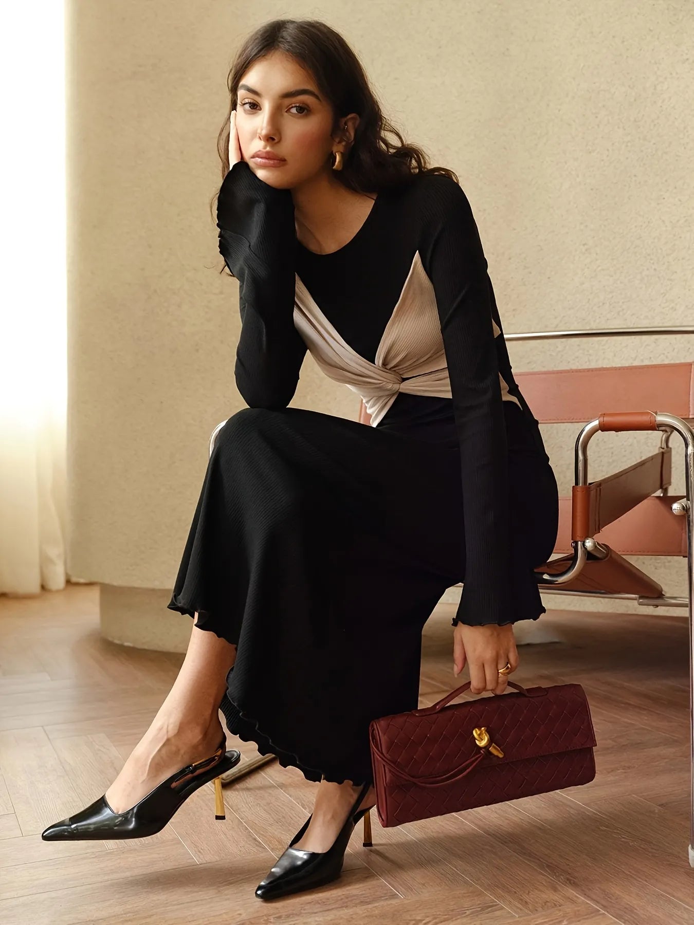 Woman wearing a long black dress with beige twist-front detail and flared sleeves. The dress has a fitted silhouette with a contrasting bodice design, styled with subtle gold hoop earrings and loose wavy hair.