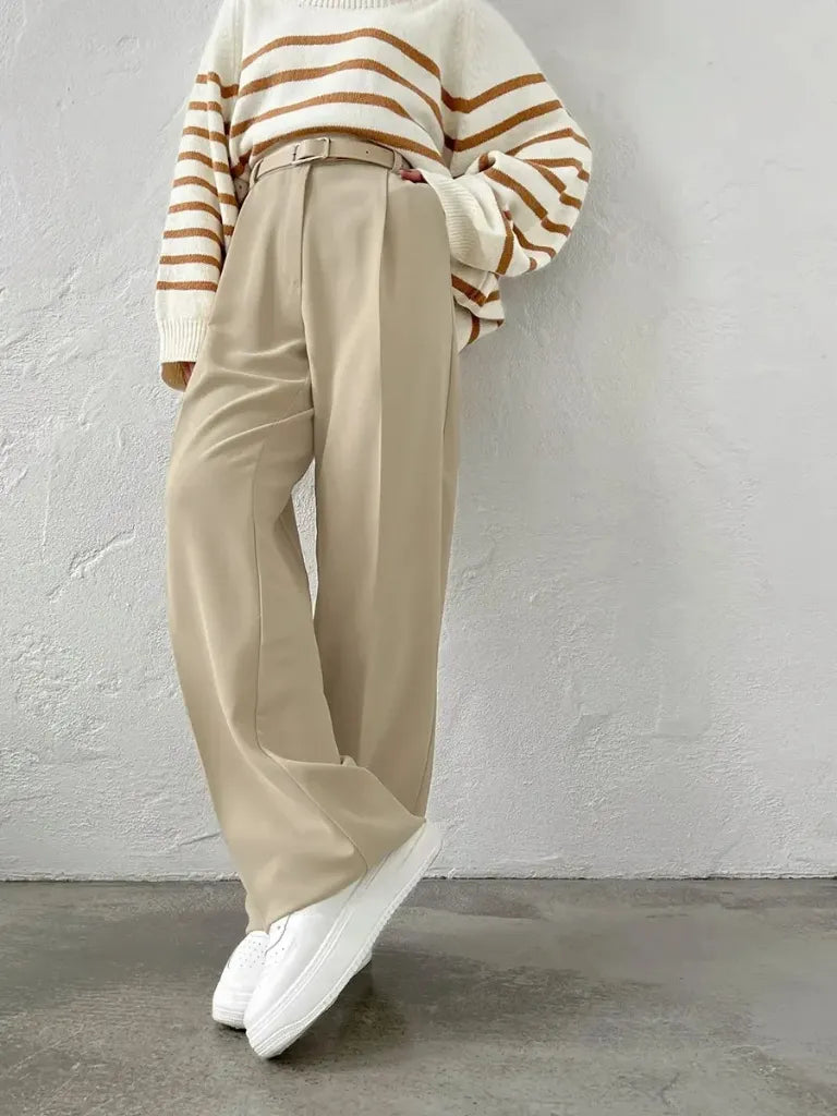Person wearing a striped sweater, beige pants, and white sneakers against a gray wall.