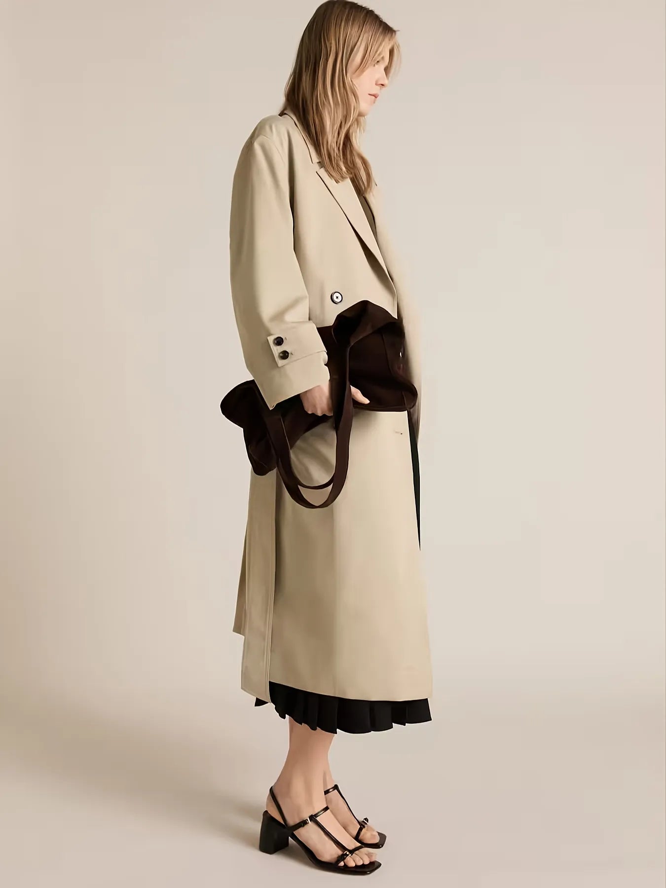 Woman wearing a beige coat with a black handbag on a plain background
