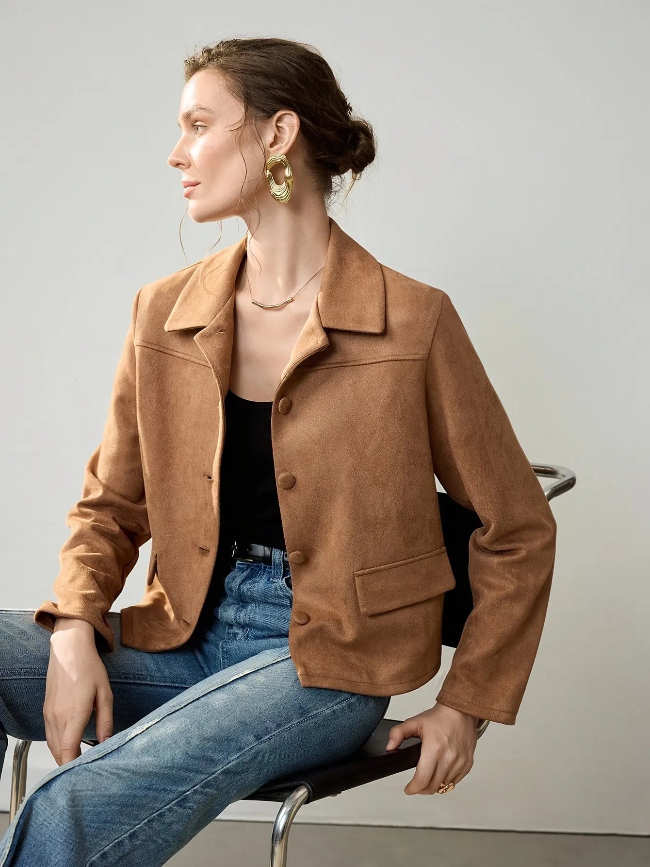 Woman wearing a cropped camel-brown suede jacket with button details, paired with a black ribbed tank top, blue jeans, and a black belt. She accessorises with large gold hoop earrings and a gold necklace, her hair styled in a loose bun.
