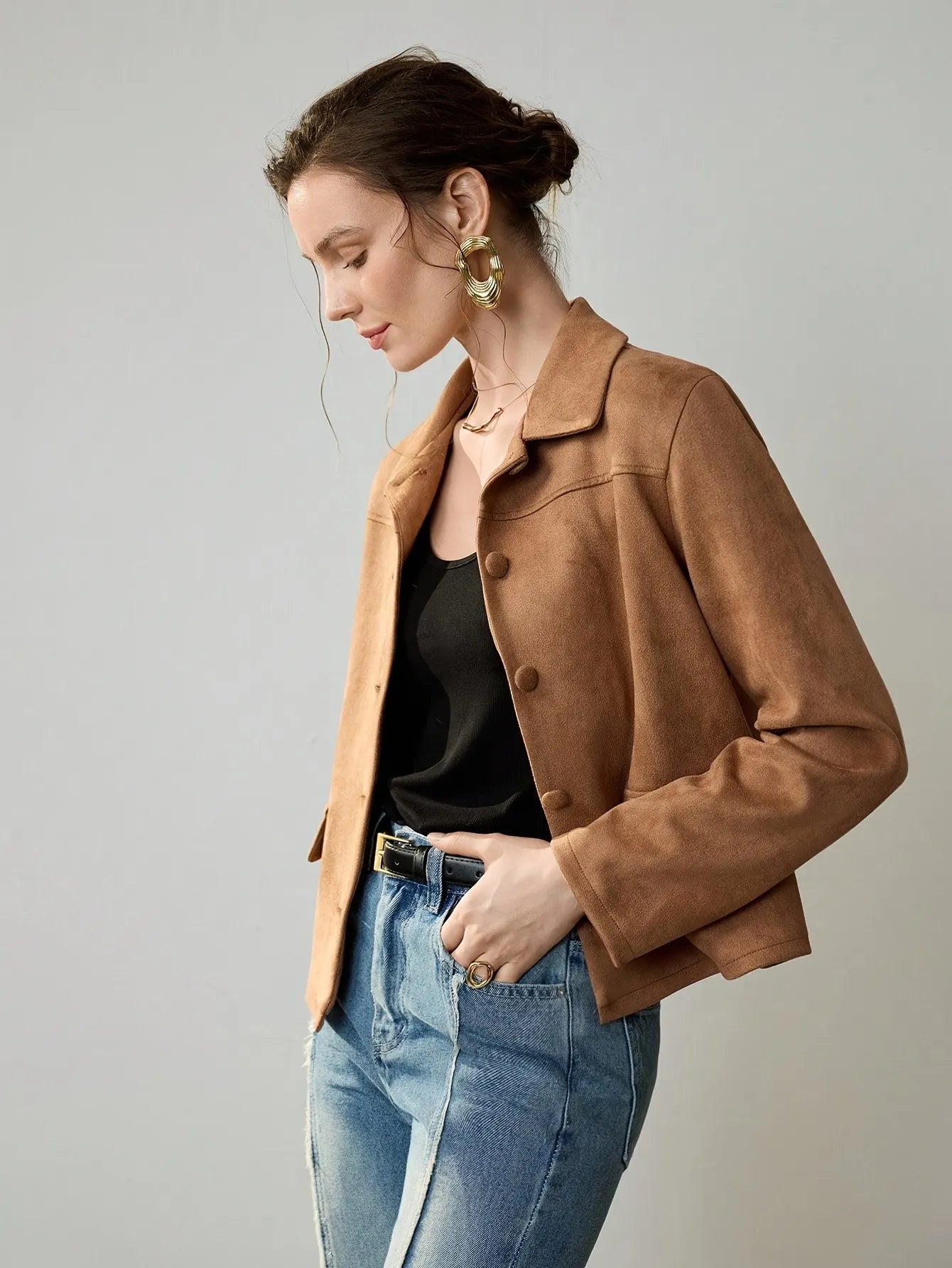Woman wearing a cropped camel-brown suede jacket with button details, paired with a black ribbed tank top, blue jeans, and a black belt. She accessorises with large gold hoop earrings and a gold necklace, her hair styled in a loose bun.