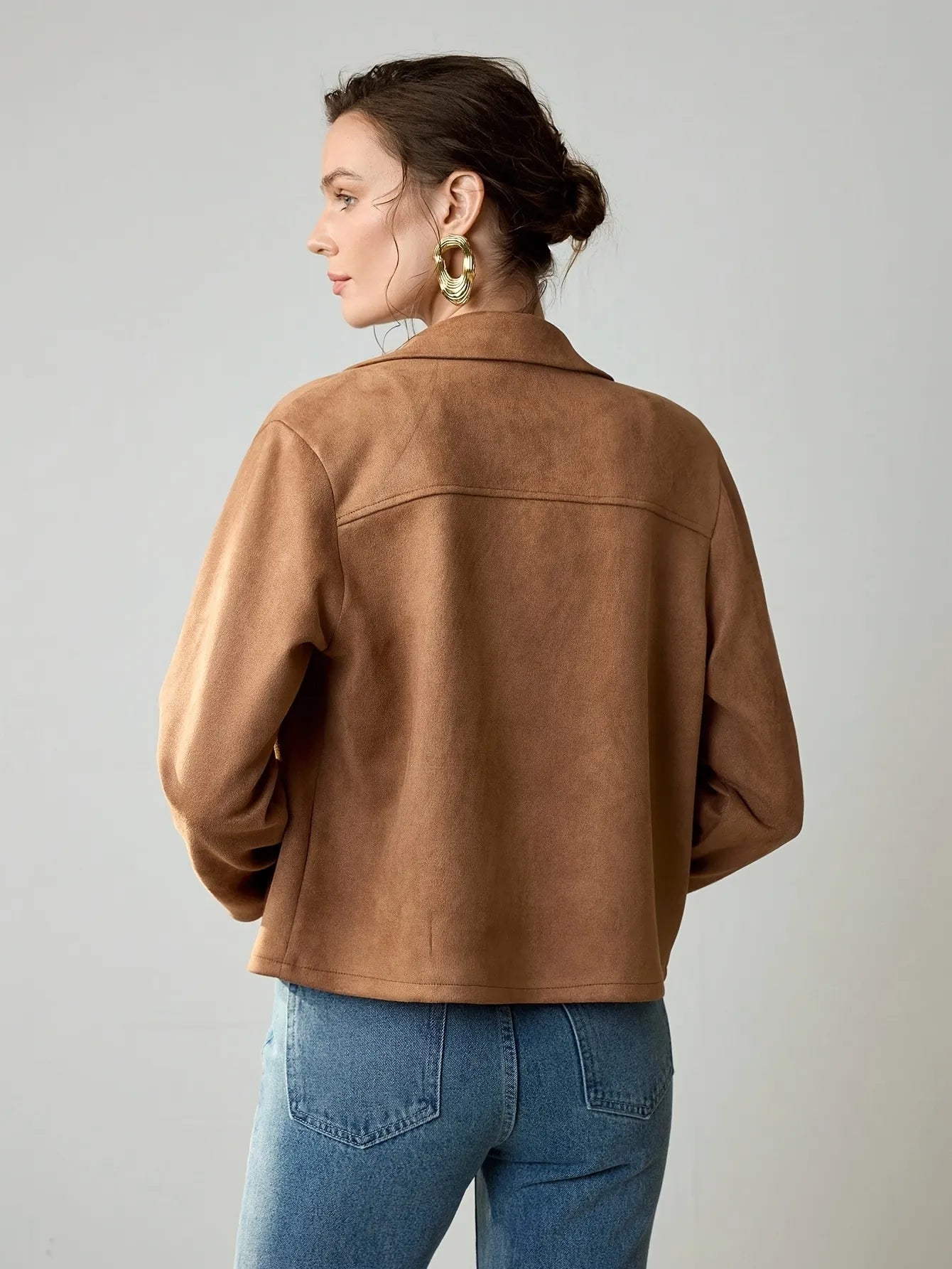 Woman wearing a cropped camel-brown suede jacket with button details, paired with a black ribbed tank top, blue jeans, and a black belt. She accessorises with large gold hoop earrings and a gold necklace, her hair styled in a loose bun.