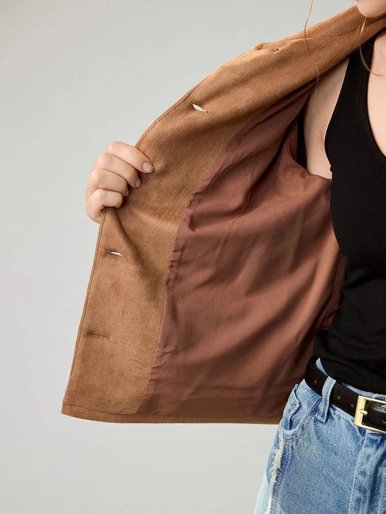 Woman wearing a cropped camel-brown suede jacket with button details, paired with a black ribbed tank top, blue jeans, and a black belt. She accessorises with large gold hoop earrings and a gold necklace, her hair styled in a loose bun.