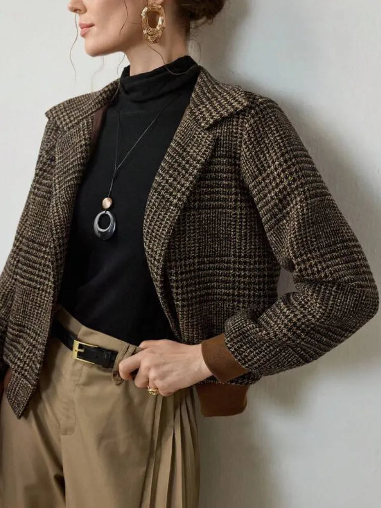 Person wearing a brown checkered blazer over a black top with beige pants against a white wall.