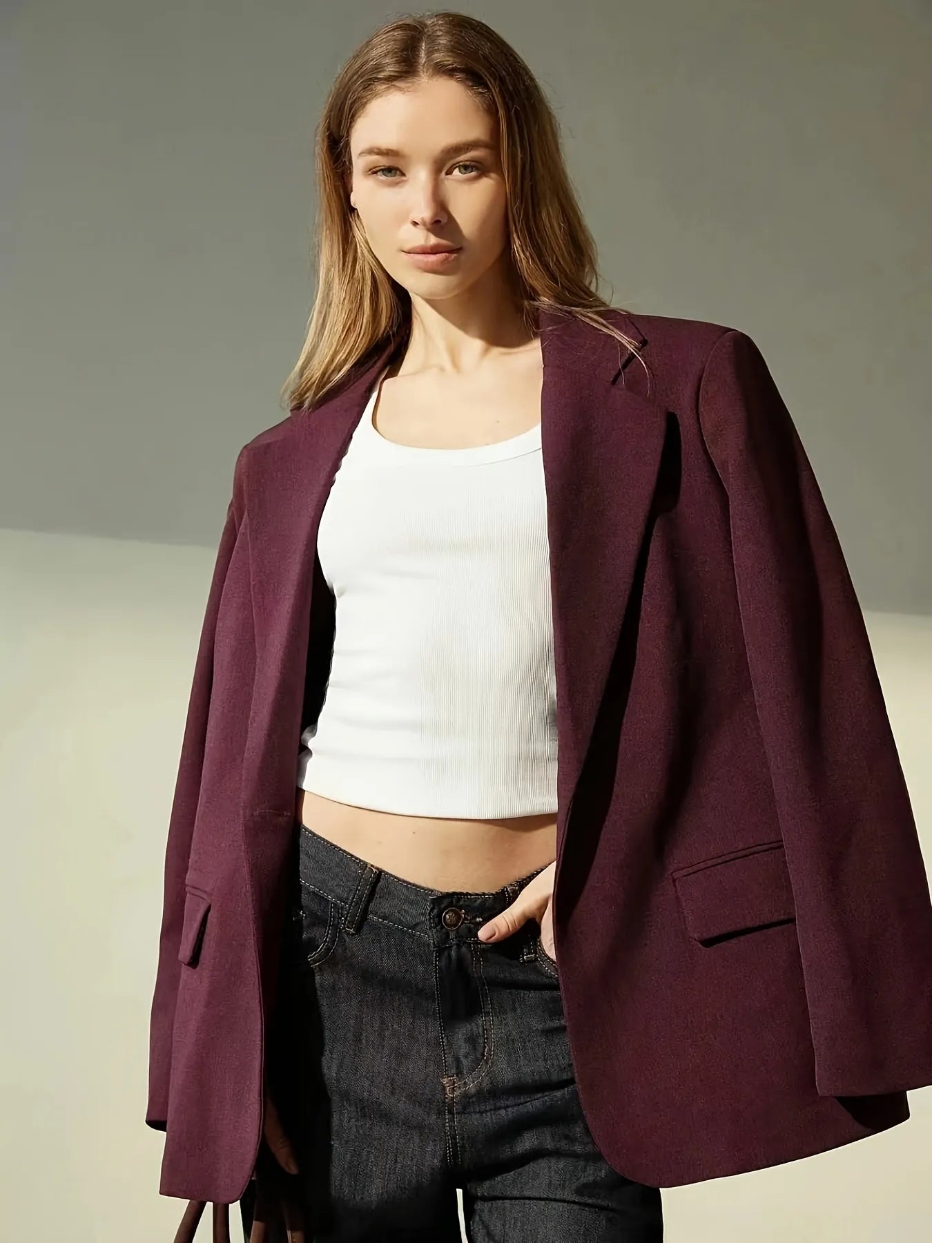 Woman wearing a burgundy blazer over a white top and dark jeans on a neutral background