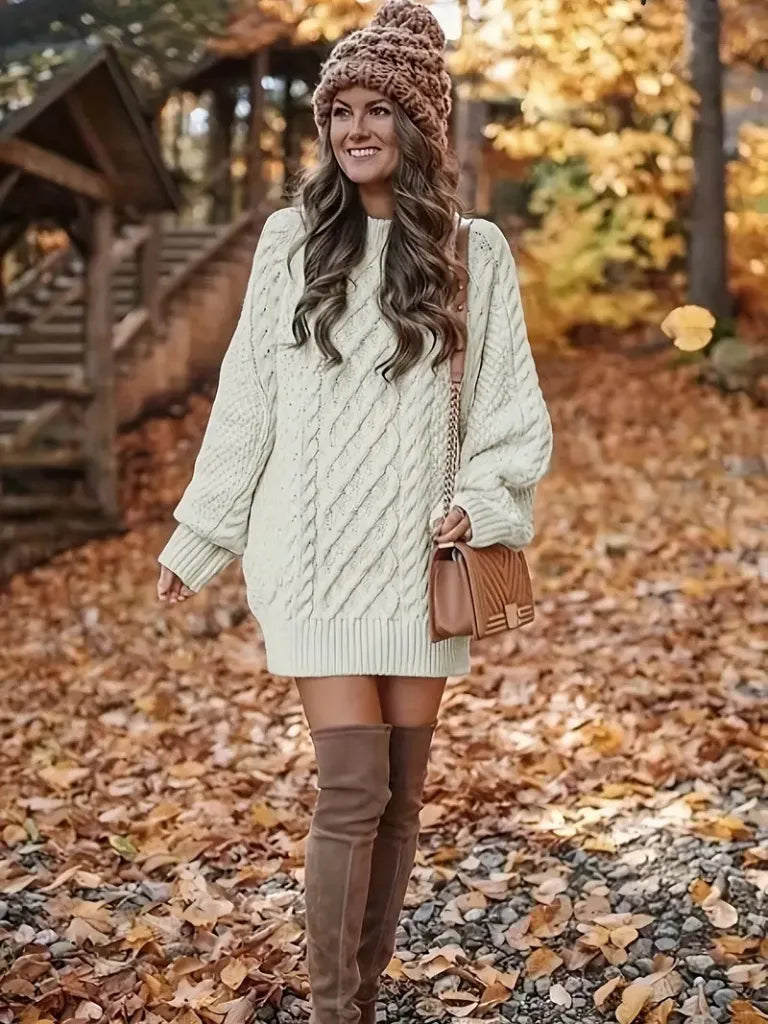 Woman in a cream cable knit sweater and brown knee-high boots standing in an autumn setting.
