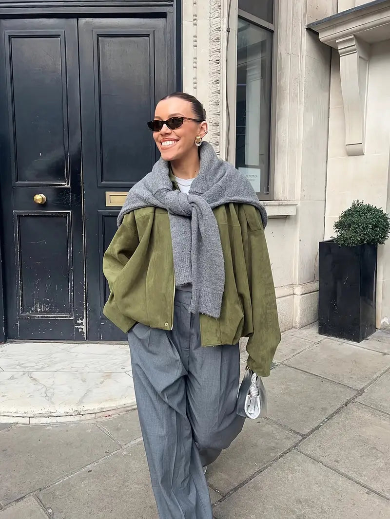 Person wearing a green coat, gray scarf, and gray pants standing on a sidewalk.