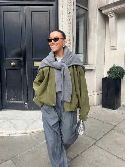 Person wearing a green coat, gray scarf, and gray pants standing on a sidewalk.