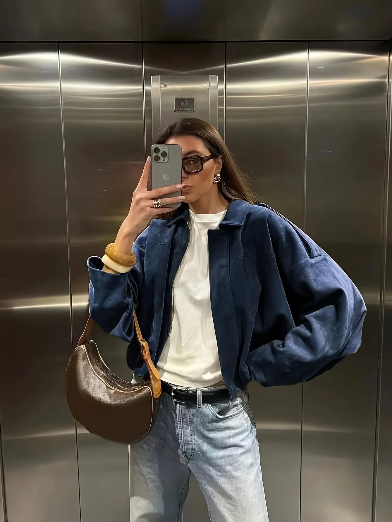 Person taking a mirror selfie wearing a blue jacket, white shirt, and jeans in an elevator.