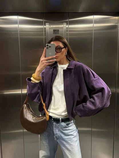 Person taking a mirror selfie wearing a purple jacket, white shirt, blue jeans, and brown bag in an elevator.