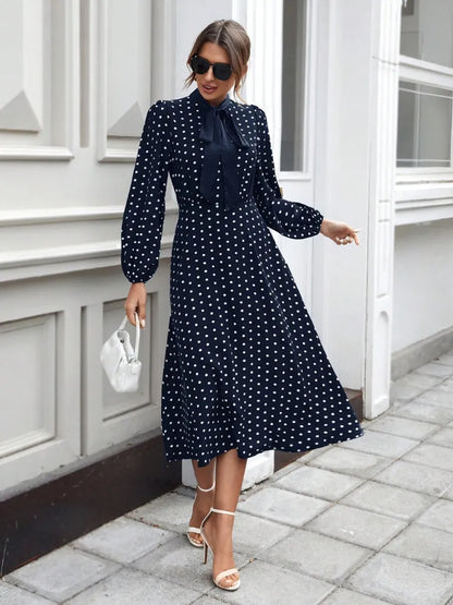 Woman wearing a navy polka dot dress with white heels and sunglasses, standing on a sidewalk.