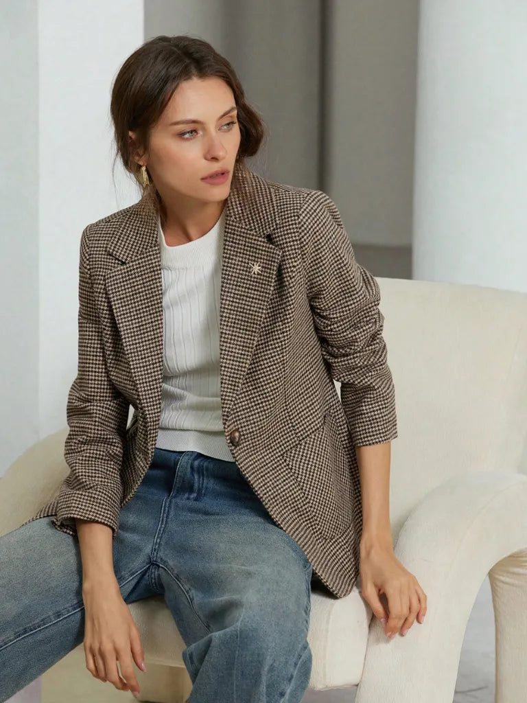 Woman sitting on a white chair wearing a brown checkered blazer and blue jeans.