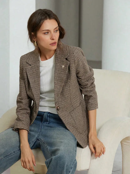 Woman sitting on a white chair wearing a brown checkered blazer and blue jeans.