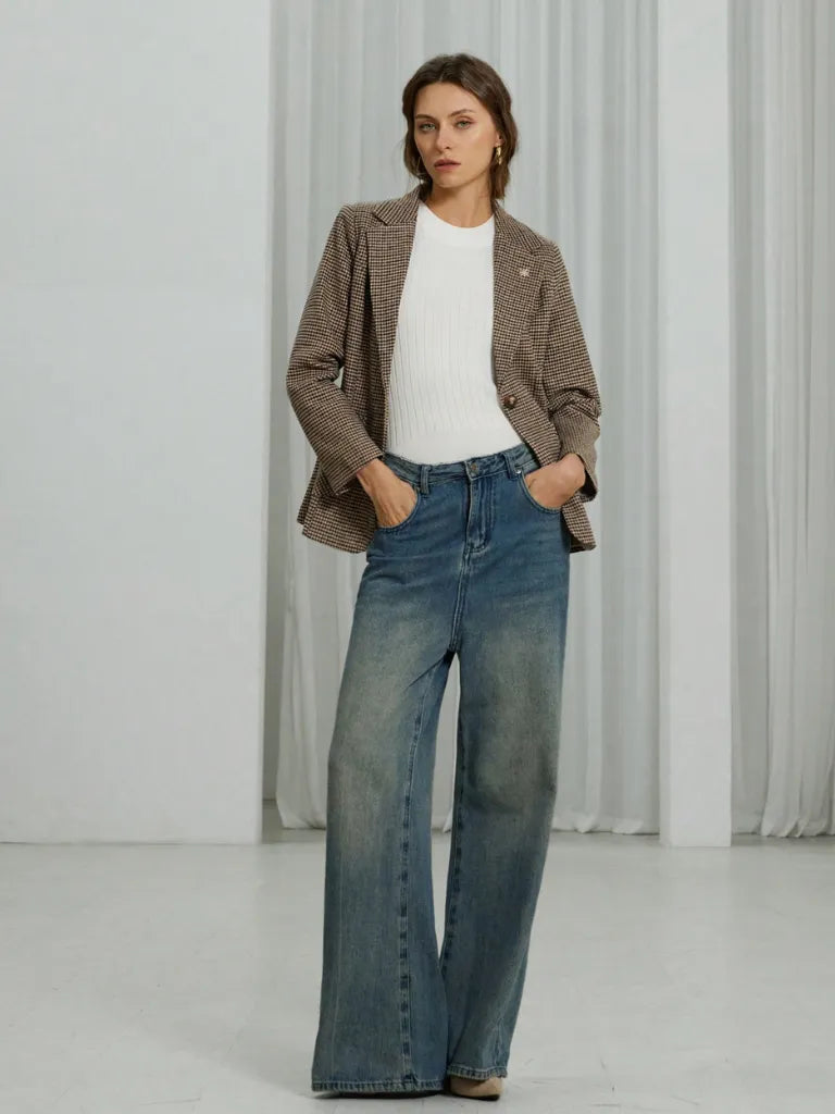 Woman wearing a brown blazer, white shirt, and blue jeans against a white curtain background