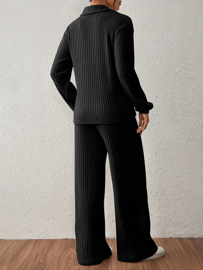 Person wearing a black ribbed sweater and pants against a white wall.