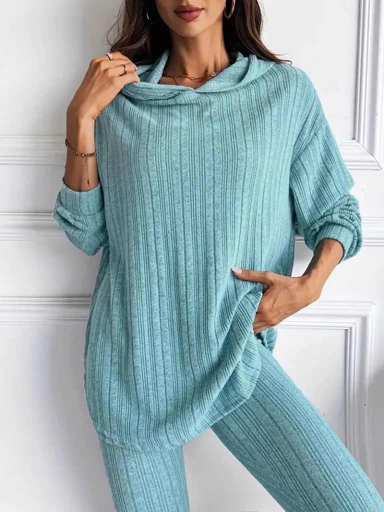 Person wearing a teal ribbed sweater and pants against a white wall
