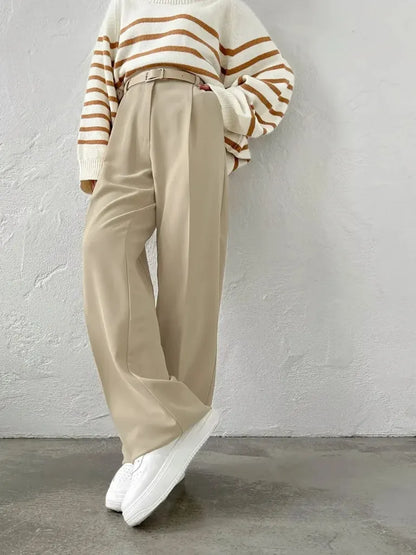 Person wearing a striped sweater, beige pants, and white sneakers against a gray wall.